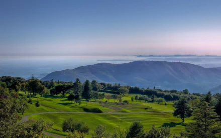 2K Quad HD PC desktop wallpaper — nature landscape of rolling green hills and scattered trees descending to misty blue mountains under a wide, clear sky.