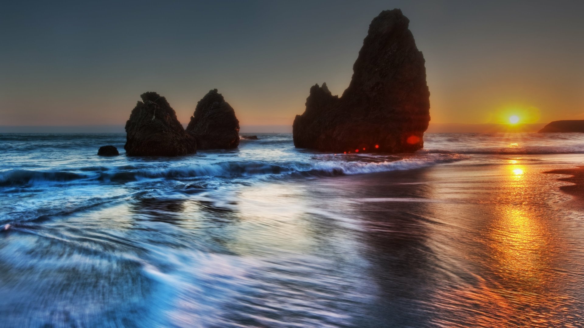 Stunning HD desktop wallpaper featuring a serene beach at sunset, with silhouetted rock formations and gentle waves reflecting the warm colors of the fading light.