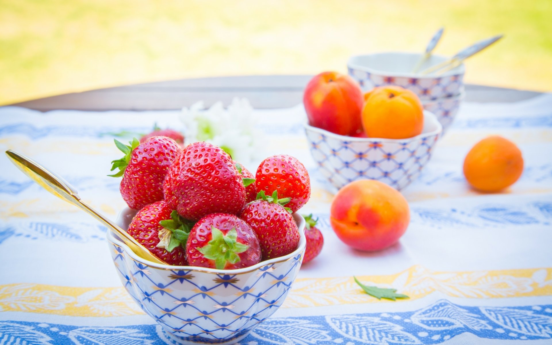 HD desktop wallpaper featuring fresh strawberries and apricots in decorative bowls on a bright, patterned tablecloth.