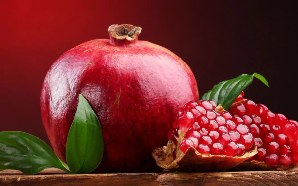 HD desktop wallpaper featuring a vibrant whole pomegranate and a half with seeds, set against a rich red background with green leaves.