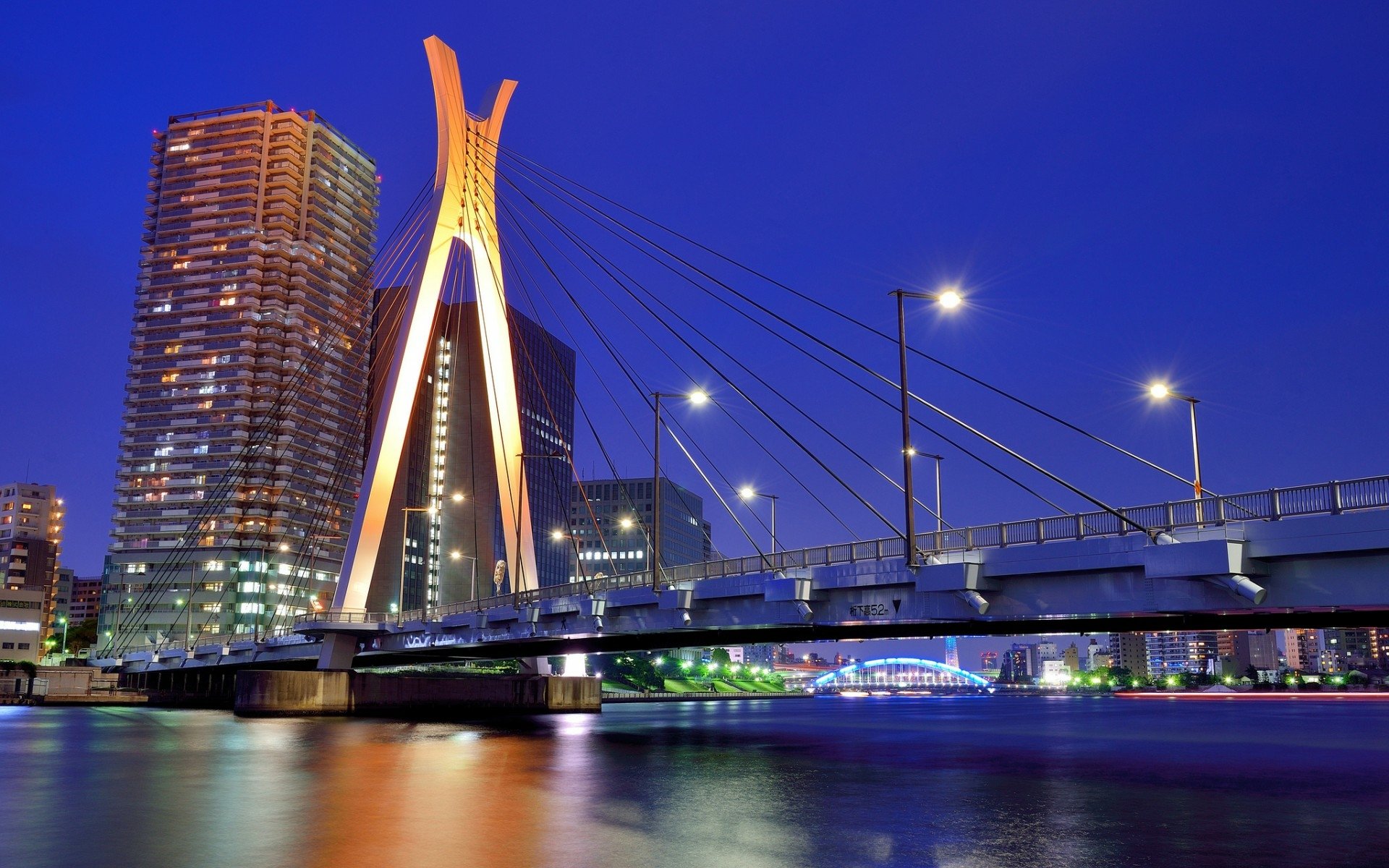 The Chuo-Ohashi Bridge illuminated against a blue night sky, reflecting over the river. Skyscrapers and vibrant lights create a stunning Tokyo metropolis scene.
