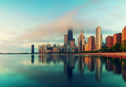 A stunning 4K Ultra HD desktop wallpaper of Chicago's skyline, showcasing tall buildings reflected in calm waters beneath a soft cloud cover.