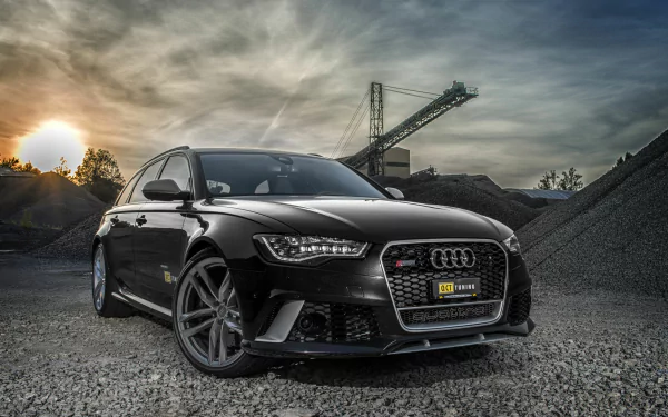 HD PC desktop wallpaper showcasing a sleek black Audi RS6 parked on gravel with an industrial backdrop under a dramatic sky at sunset.