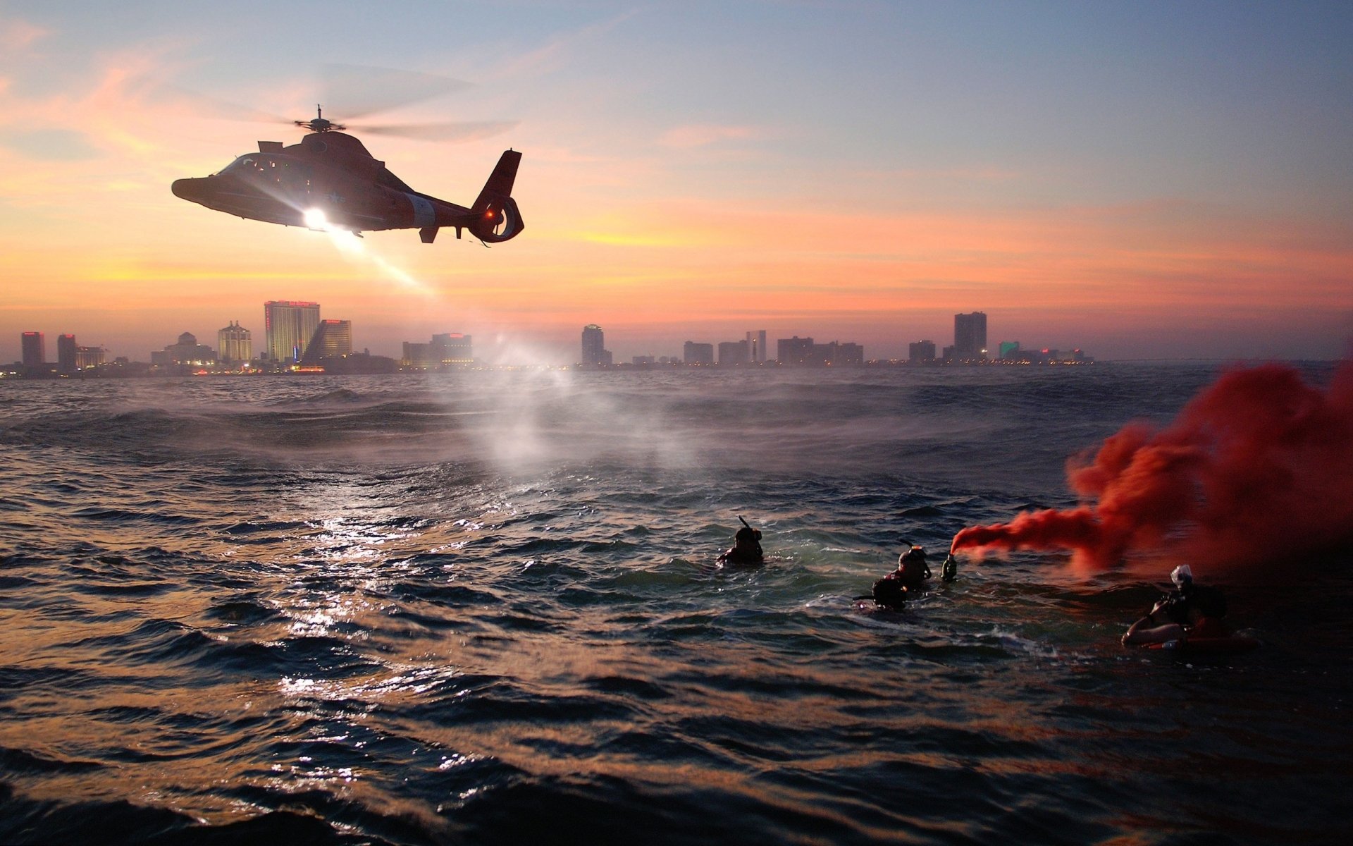A helicopter hovers above water, with rescue divers below and a vibrant sunset casting colors over a city skyline, creating a dramatic scene for an HD desktop wallpaper.