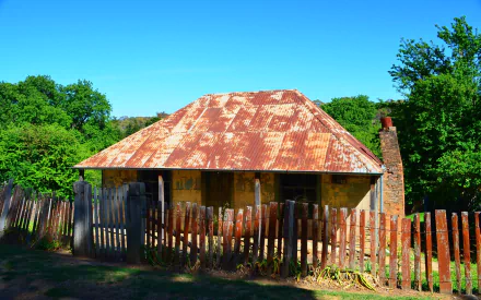 Historic Beyers Cottage — old man-made cottage building with rusted corrugated roof and weathered wooden fence amid green trees; HD PC desktop wallpaper background.