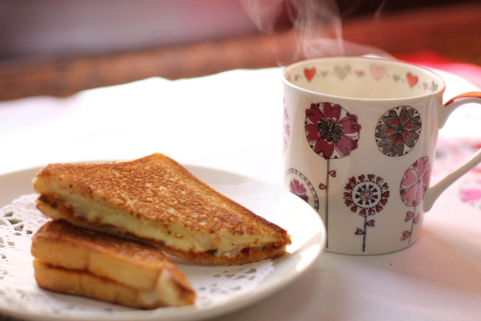 HD PC desktop wallpaper of food, sandwich: toasted grilled sandwich on a doily-lined plate beside a steaming floral mug, soft-lit cozy breakfast scene.