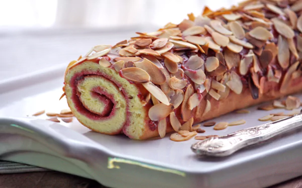 A beautifully presented cake roll adorned with almond slices, resting on a white platter. This 4K Ultra HD wallpaper showcases delicious food in vibrant detail.