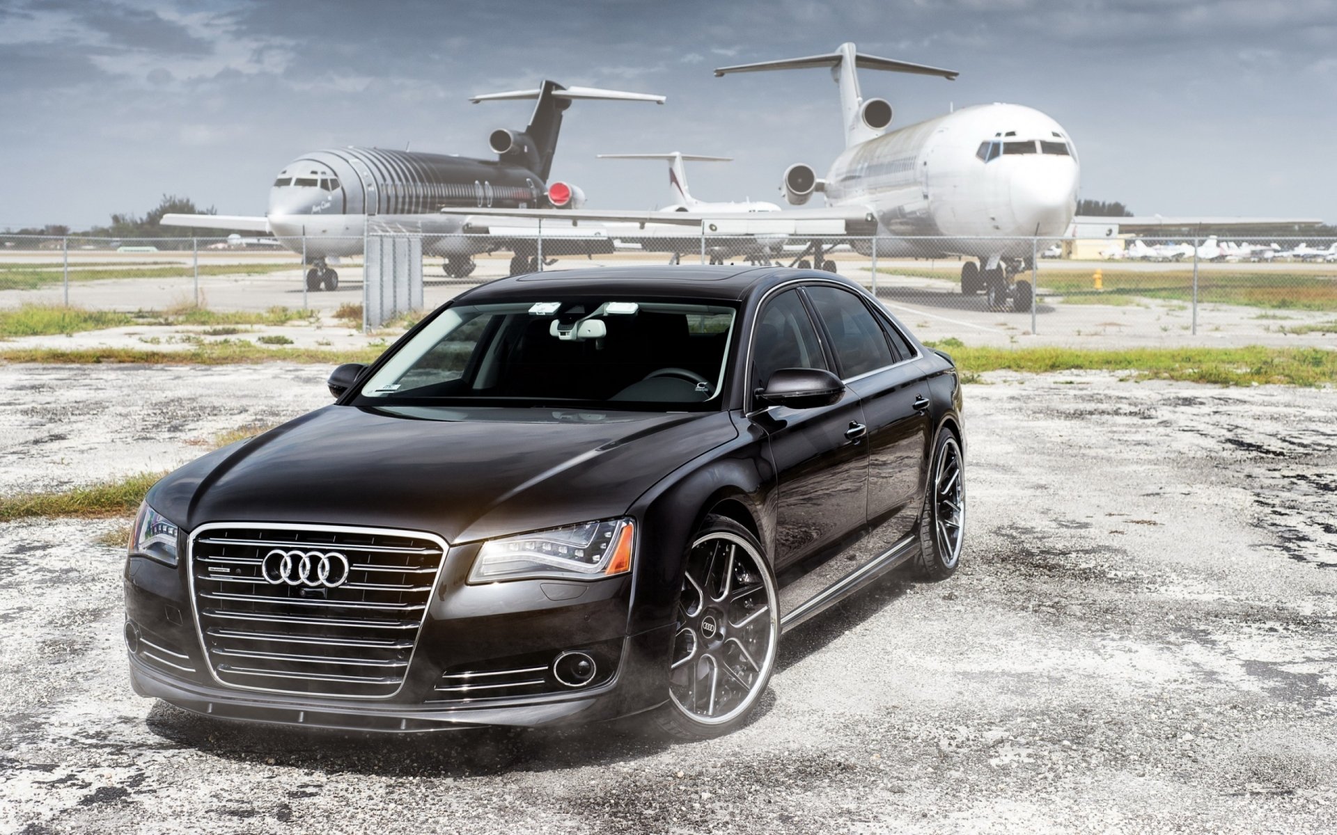 A sleek Audi A8 parked on an airstrip, with aircraft in the background, showcased in a high-definition desktop wallpaper setting.