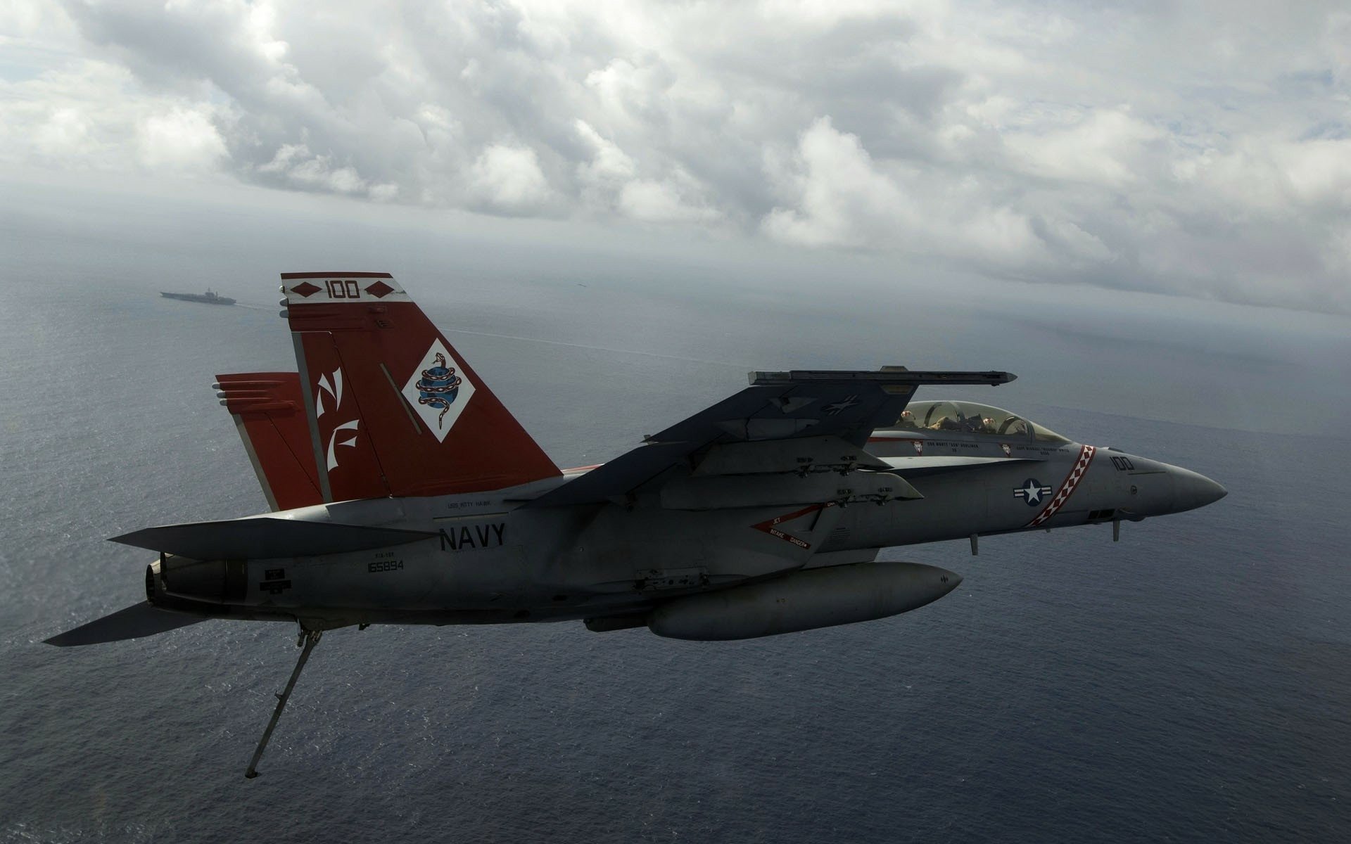 Military Boeing F/A-18E/F Super Hornet flying over the ocean with an aircraft carrier on the horizon under cloudy skies, HD PC desktop wallpaper and background.