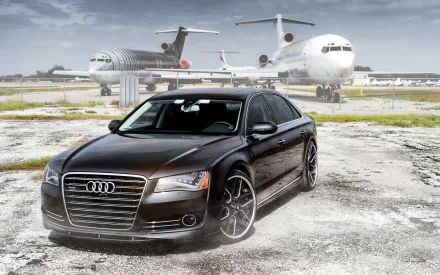 A sleek Audi A8 parked on an airstrip, with aircraft in the background, showcased in a high-definition desktop wallpaper setting.