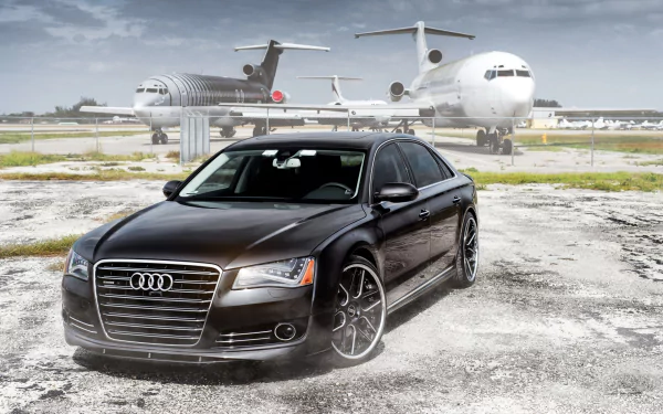 A sleek Audi A8 parked on an airstrip, with aircraft in the background, showcased in a high-definition desktop wallpaper setting.
