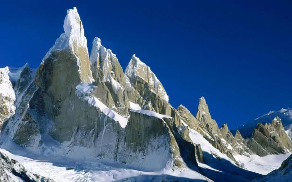 Snow-covered jagged cliffs rise sharply under a clear blue sky at sunrise in Los Glaciares National Park, Argentina, showcasing a stunning snowy mountain peak.