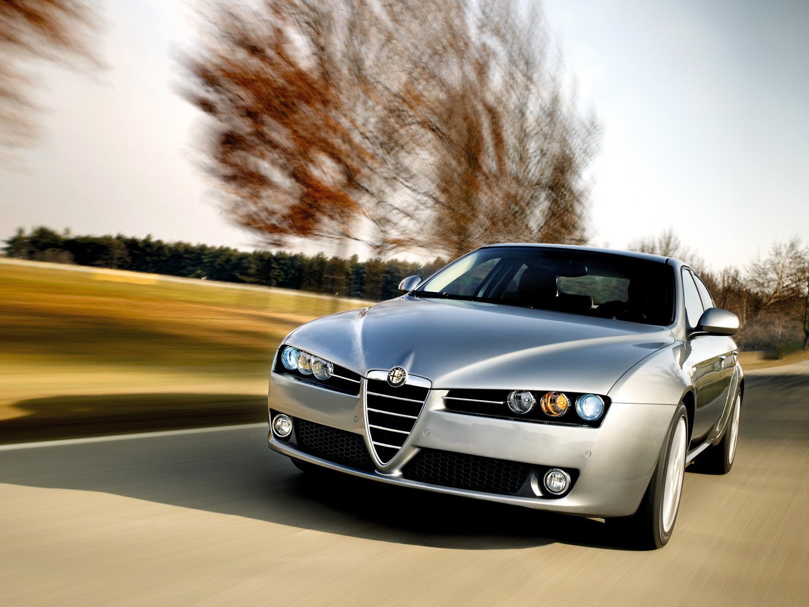 HD PC desktop wallpaper background: silver Alfa Romeo 159 vehicle speeding on a sunlit road, front three-quarter view with motion-blurred landscape.
