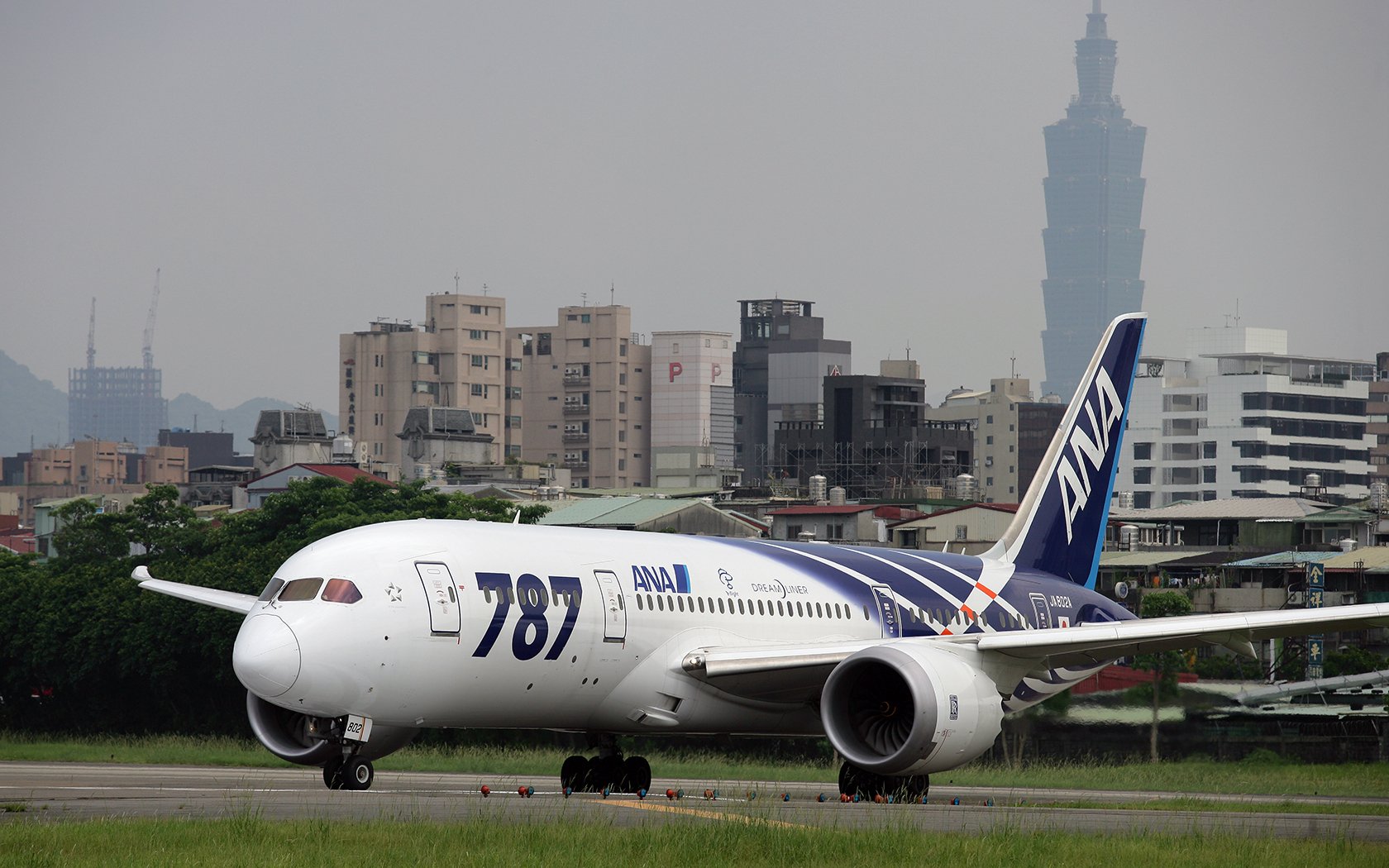 HD desktop wallpaper featuring a Boeing 787 Dreamliner airplane from ANA taxiing on a runway with city buildings and Taipei 101 tower in the background.