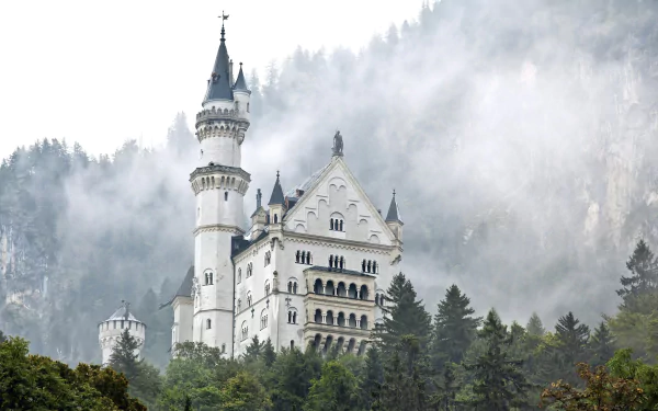 HD wallpaper of Neuschwanstein Castle, nestled among misty, forested mountains. This captivating man-made structure stands majestically, surrounded by lush greenery and low-hanging clouds.