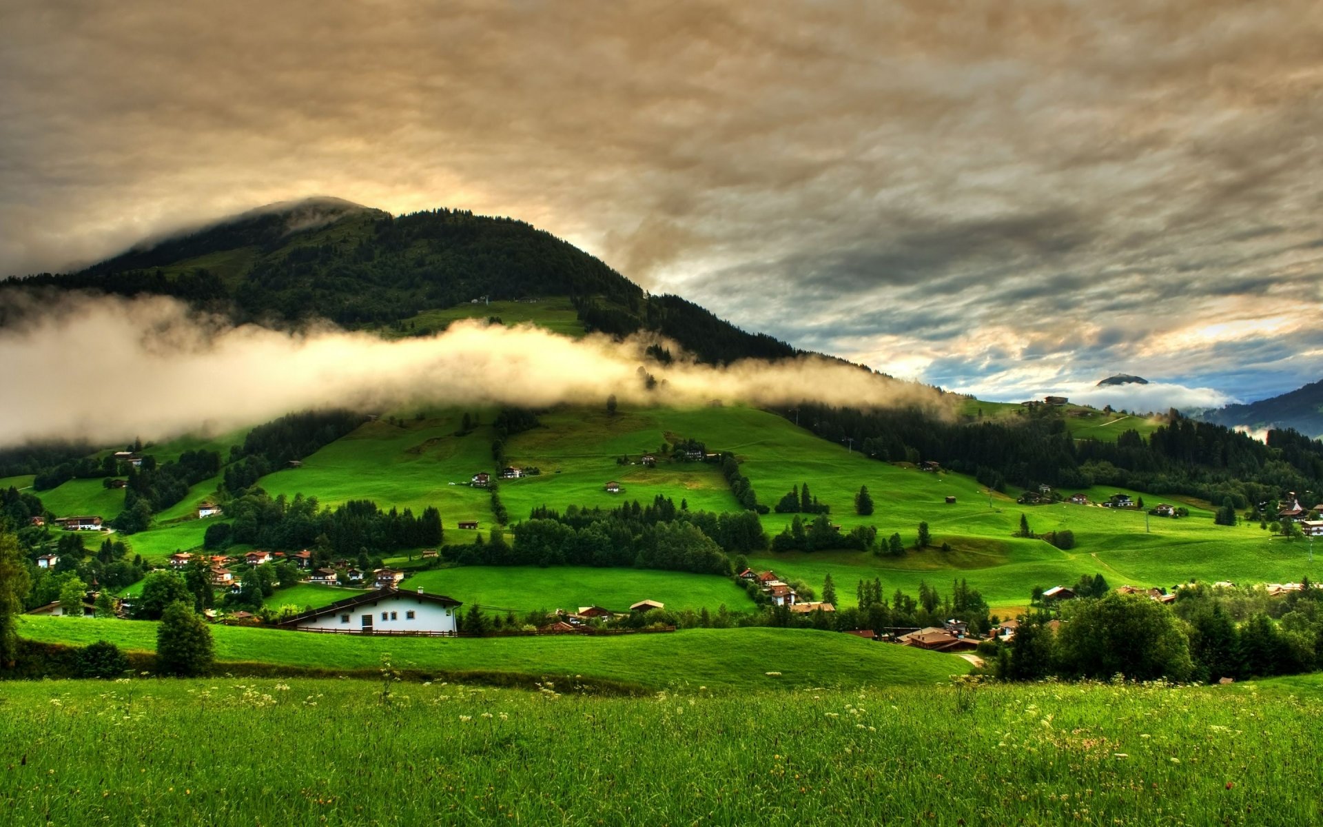 HD landscape photography of a green valley with scattered houses, low-hanging clouds, and a forested mountain under a dramatic sky, used as a PC desktop wallpaper.