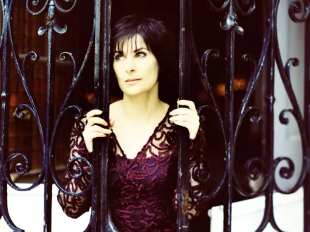 HD music desktop wallpaper: contemplative female singer behind ornate wrought-iron bars, wearing deep plum lace, moody Enya-inspired atmosphere.
