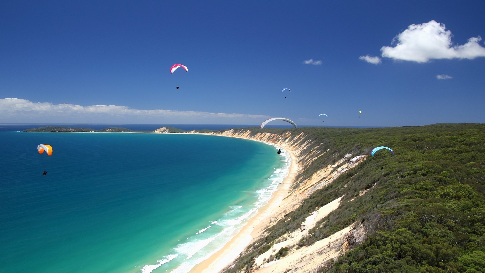 Paragliding over a curved turquoise bay with sandy cliffs and green coastal scrub, vibrant sports scene — 4K Ultra HD PC desktop wallpaper and background.