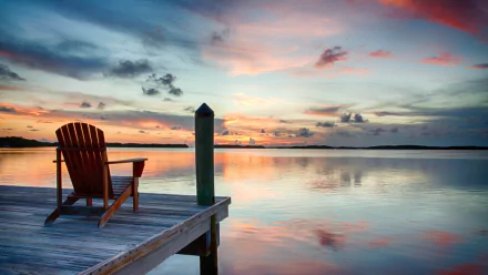 HD desktop wallpaper of a serene lake at sunset, featuring a wooden chair on a dock under a colorful sky with reflections on the calm water.