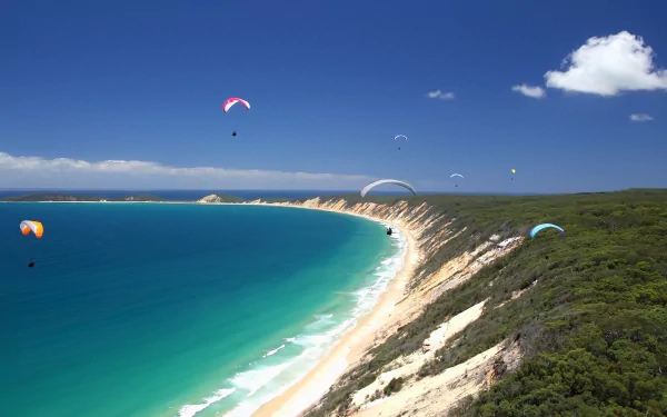 Paragliding over a curved turquoise bay with sandy cliffs and green coastal scrub, vibrant sports scene — 4K Ultra HD PC desktop wallpaper and background.