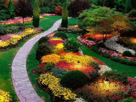 A beautiful HD desktop wallpaper of a man-made garden featuring vibrant flower beds, neatly manicured lawns, and a winding stone path through colorful, lush greenery.