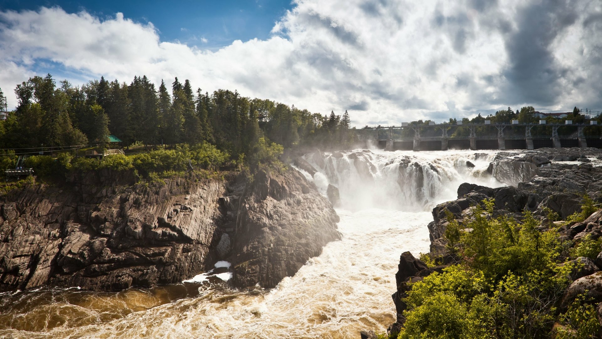 4K Ultra HD PC desktop wallpaper and background showing a nature scene: a powerful waterfall cascading through rocky cliffs into a turbulent river, framed by pine forest and dramatic sky.