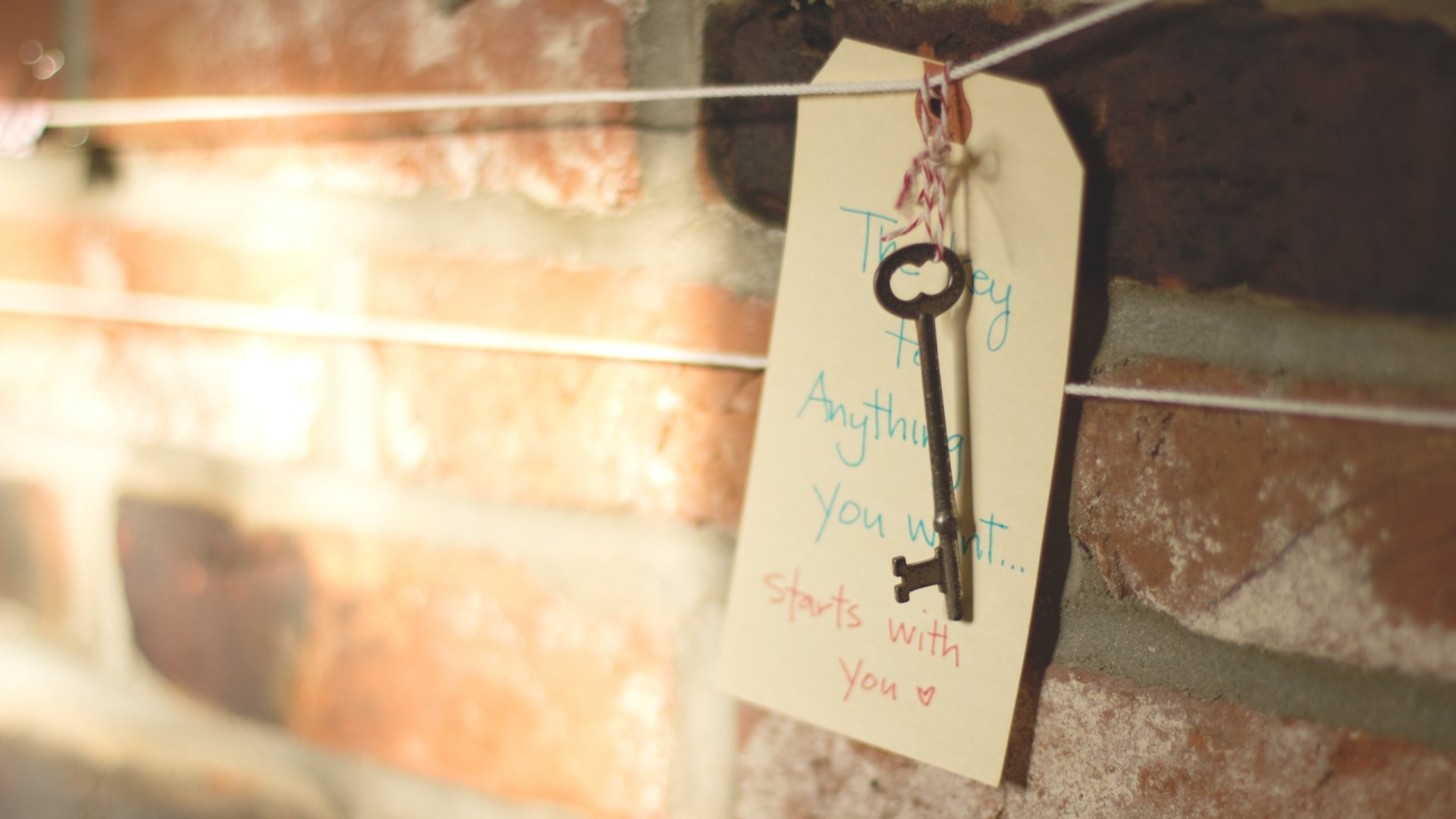 HD desktop wallpaper featuring a close-up of a man-made vintage key hanging on a string against a rustic brick wall, with a handwritten tag attached.