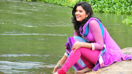 Brunette Bollywood celebrity Deeksha Seth in vibrant traditional attire smiles by a serene riverside, captured in 4K Ultra HD PC desktop wallpaper quality.