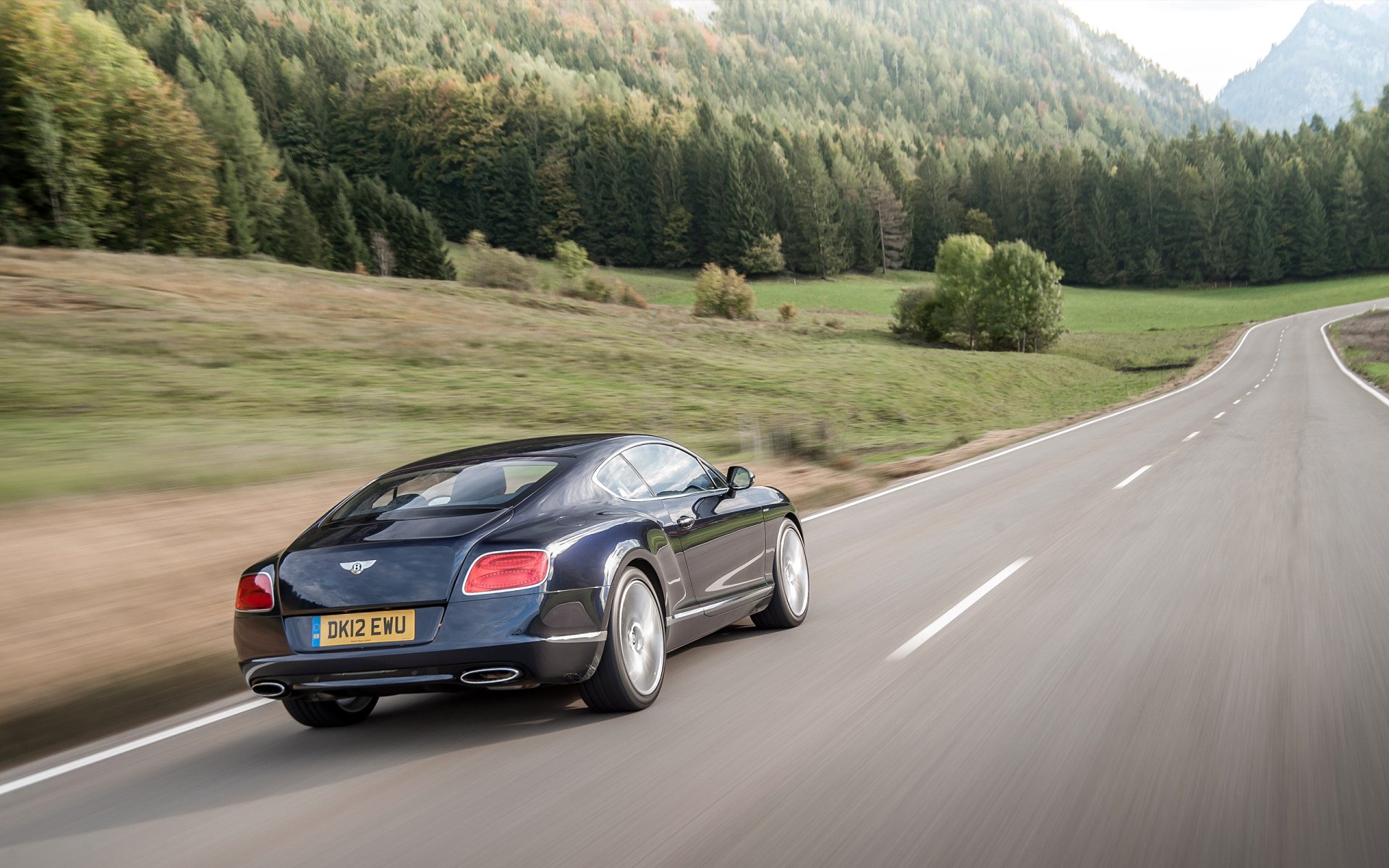 A black Bentley Continental GT Speed driving on a winding road through a scenic forested landscape, captured in an HD desktop wallpaper image.