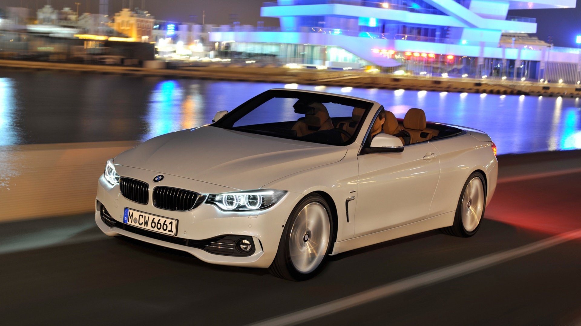 HD PC desktop wallpaper background of a white BMW 4 Series Cabrio vehicle driving at night by a waterfront, city lights reflecting on the water.