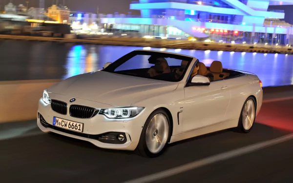 HD PC desktop wallpaper background of a white BMW 4 Series Cabrio vehicle driving at night by a waterfront, city lights reflecting on the water.