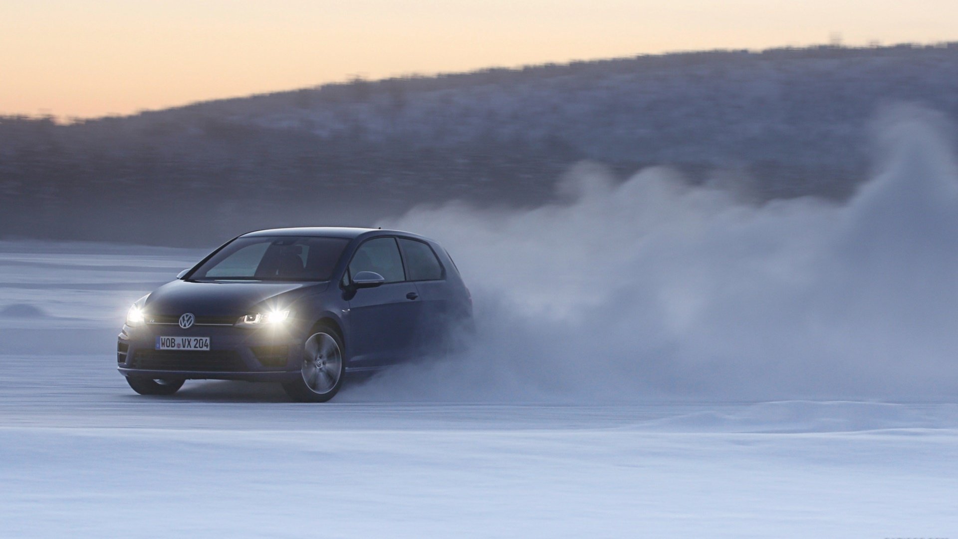 HD PC desktop wallpaper featuring a Volkswagen Golf R drifting on a snowy landscape with headlights on against a backdrop of hills at sunset.