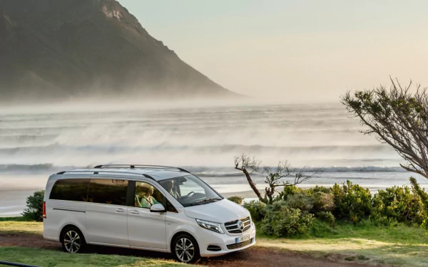 HD PC desktop wallpaper/background: white Mercedes‑Benz V‑Class parked on a coastal path with misty waves, a rugged mountain backdrop and coastal shrubs under a soft sky.