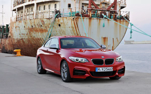 A red BMW M235i Coupe parked on a dock with a rusted ship in the background, captured in an HD PC desktop wallpaper style.