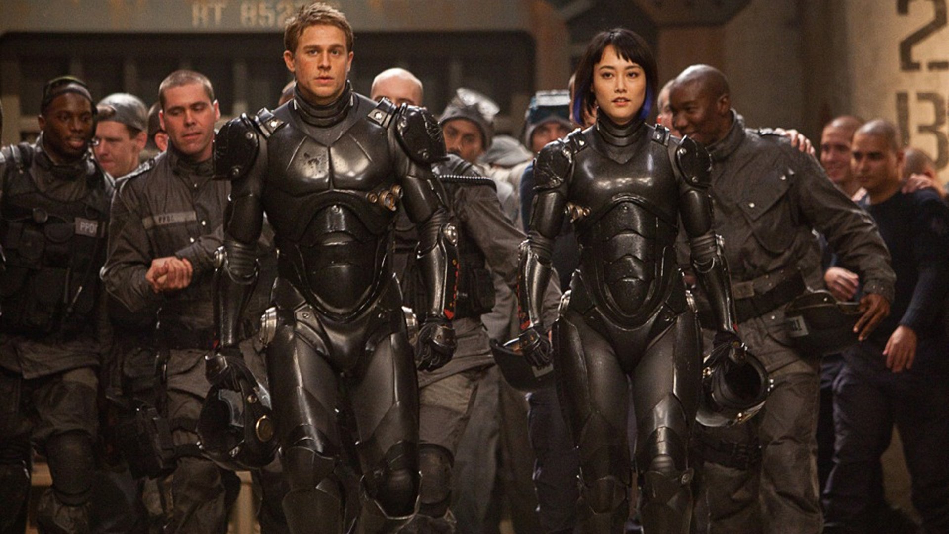 Rinko Kikuchi and Charlie Hunnam in full combat suits walk confidently among a group, from the movie Pacific Rim, featured in an HD PC desktop wallpaper.
