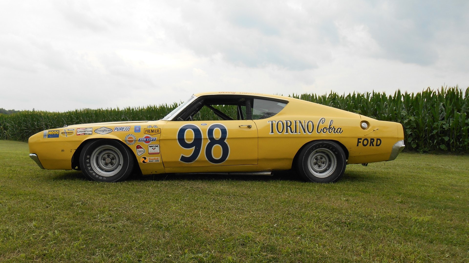 Yellow Ford Torino Cobra race car (#98) side view on grass beneath a cloudy sky — 5K Ultra HD PC desktop wallpaper and background.
