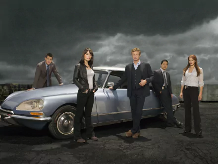 A dramatic scene from *The Mentalist* featuring main characters Patrick Jane, Teresa Lisbon, Wayne Rigsby, Kimball Cho, and Grace Van Pelt standing beside a classic car against a moody sky.
