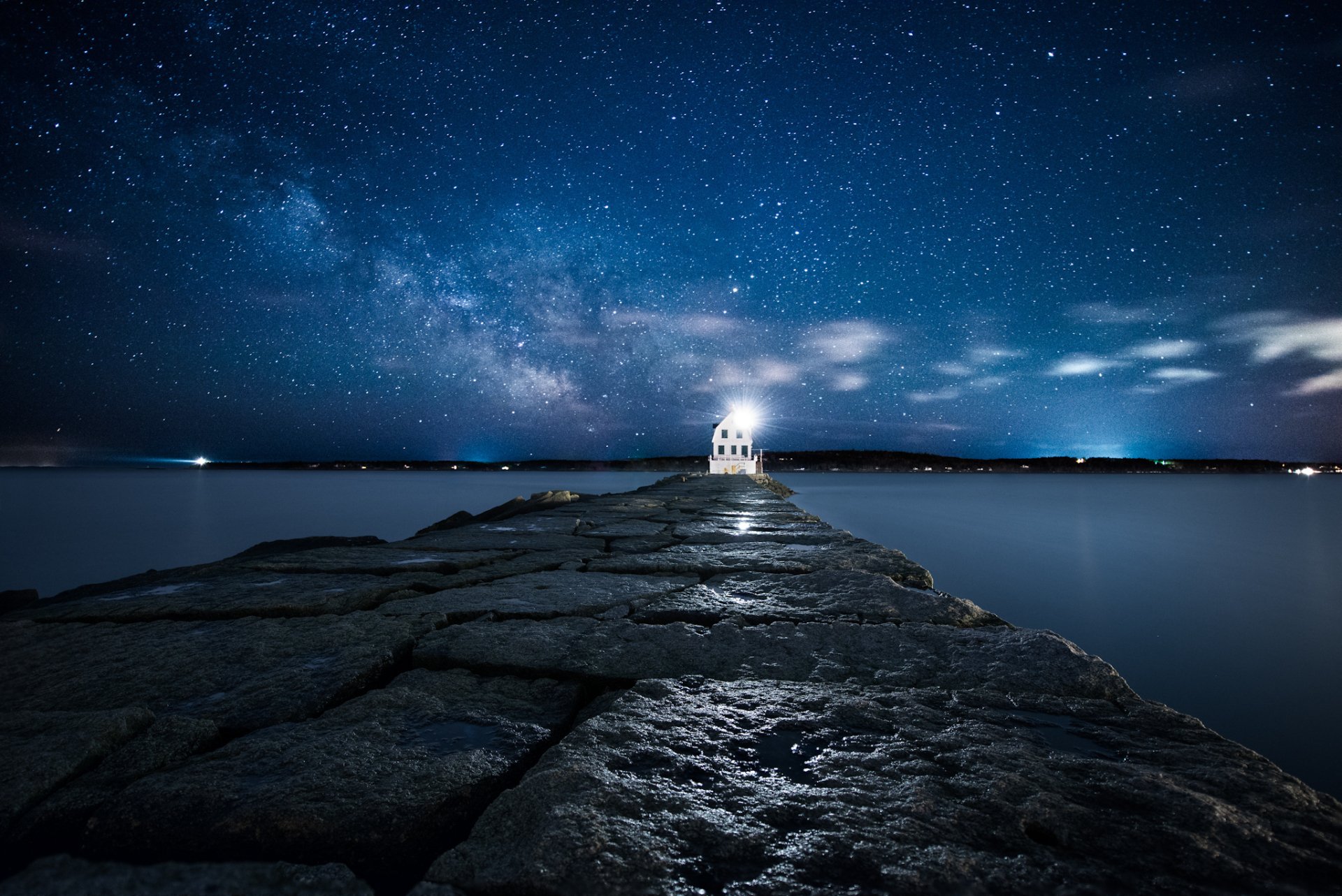 A serene night scene featuring a lighthouse illuminating a rocky pier under a starry sky, creating a stunning HD desktop wallpaper and background.