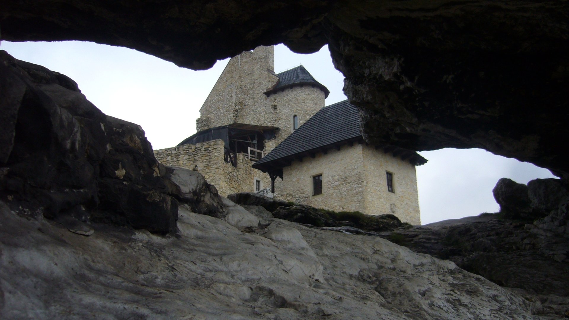 Man-made Bobolice Castle seen through a rocky cave opening, stone towers against a pale sky — 2K Quad HD PC desktop wallpaper/background.