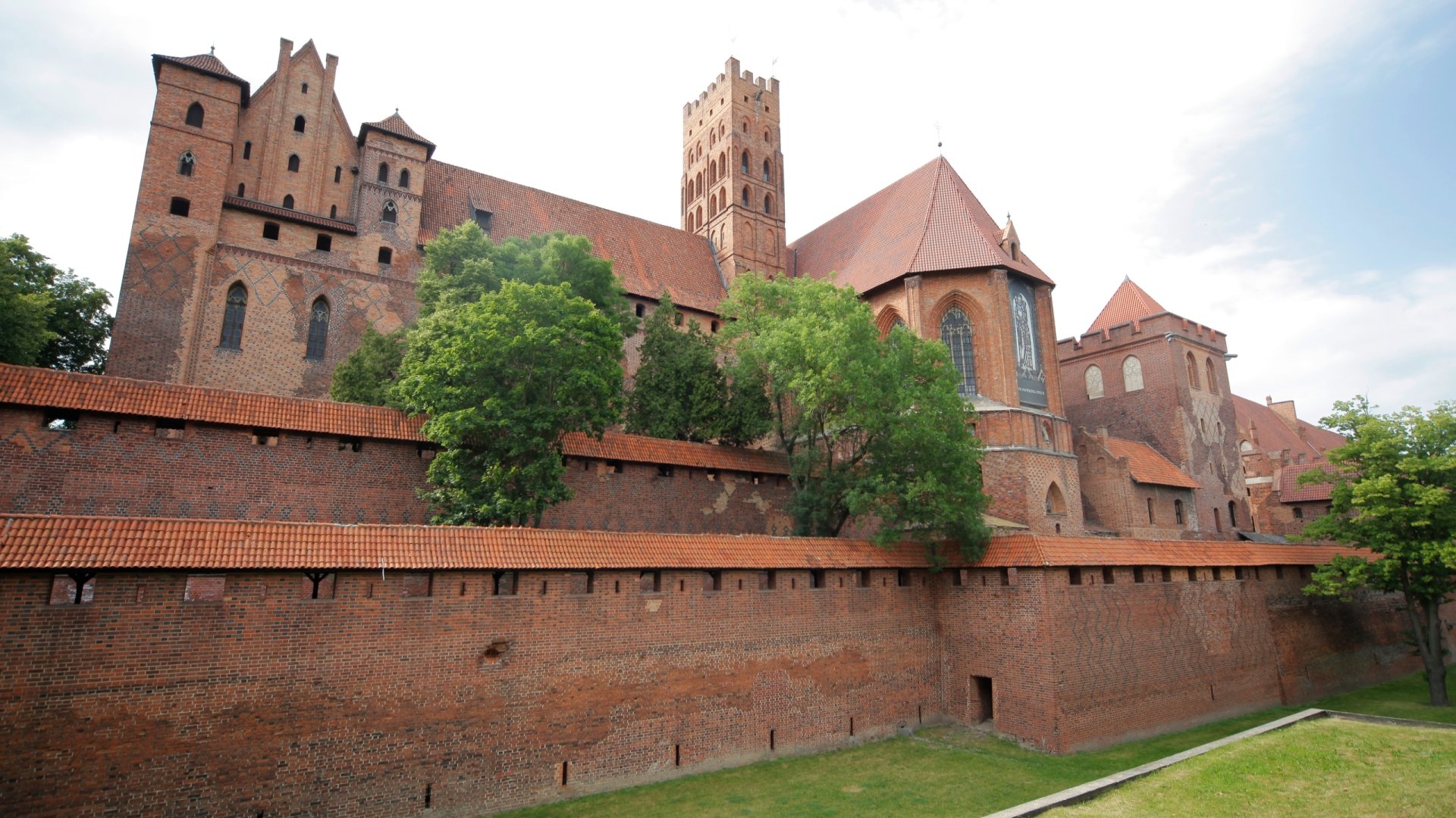 Malbork Castle, a sprawling red-brick medieval fortress with fortified walls and trees, shown as a 2K Quad HD PC desktop wallpaper background — man-made architecture