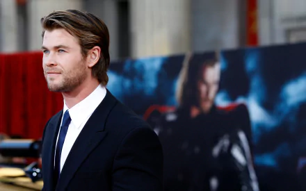 HD desktop wallpaper featuring Australian actor and celebrity Chris Hemsworth dressed in a black suit at a red carpet event with a blurred Thor poster in the background.