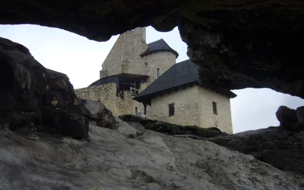 Man-made Bobolice Castle seen through a rocky cave opening, stone towers against a pale sky — 2K Quad HD PC desktop wallpaper/background.