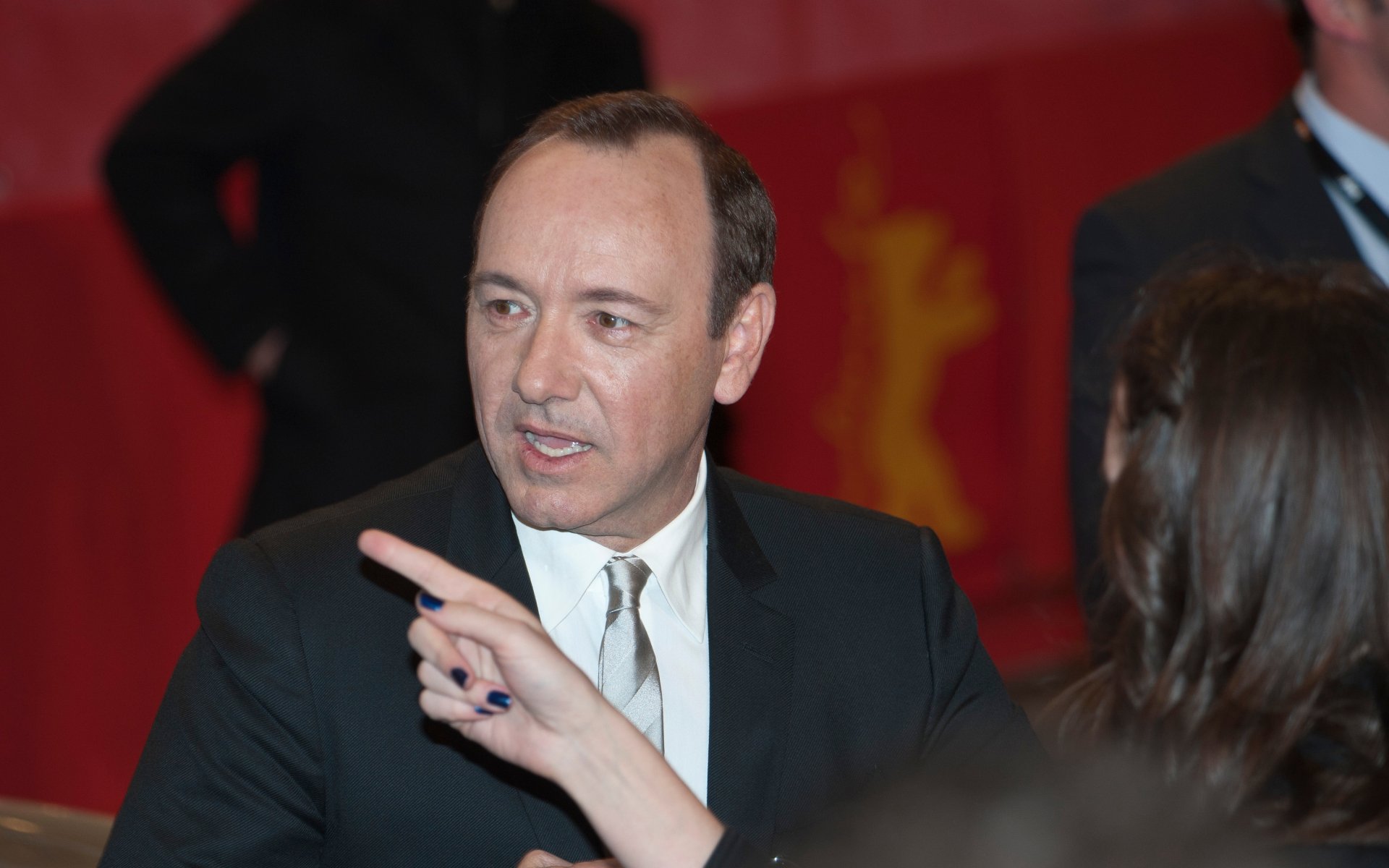 Man in dark suit and tie at a red-carpet event, speaking and pointing with a blurred crowd behind — 2K Quad HD PC desktop wallpaper.