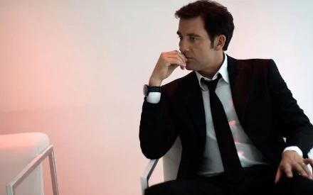 HD PC desktop wallpaper featuring English actor Clive Owen dressed in a black suit, sitting thoughtfully against a minimalist light background.