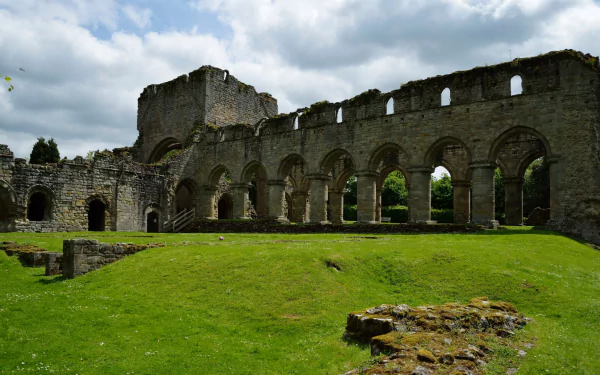 HD PC desktop wallpaper: religious ruins of Buildwas Abbey — medieval stone arches over a green lawn under a dramatic cloudy sky.