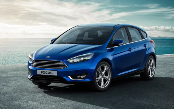 A blue 2015 Ford Focus hatchback parked on a coastal road under a cloudy sky, featured as an HD PC desktop wallpaper and background.