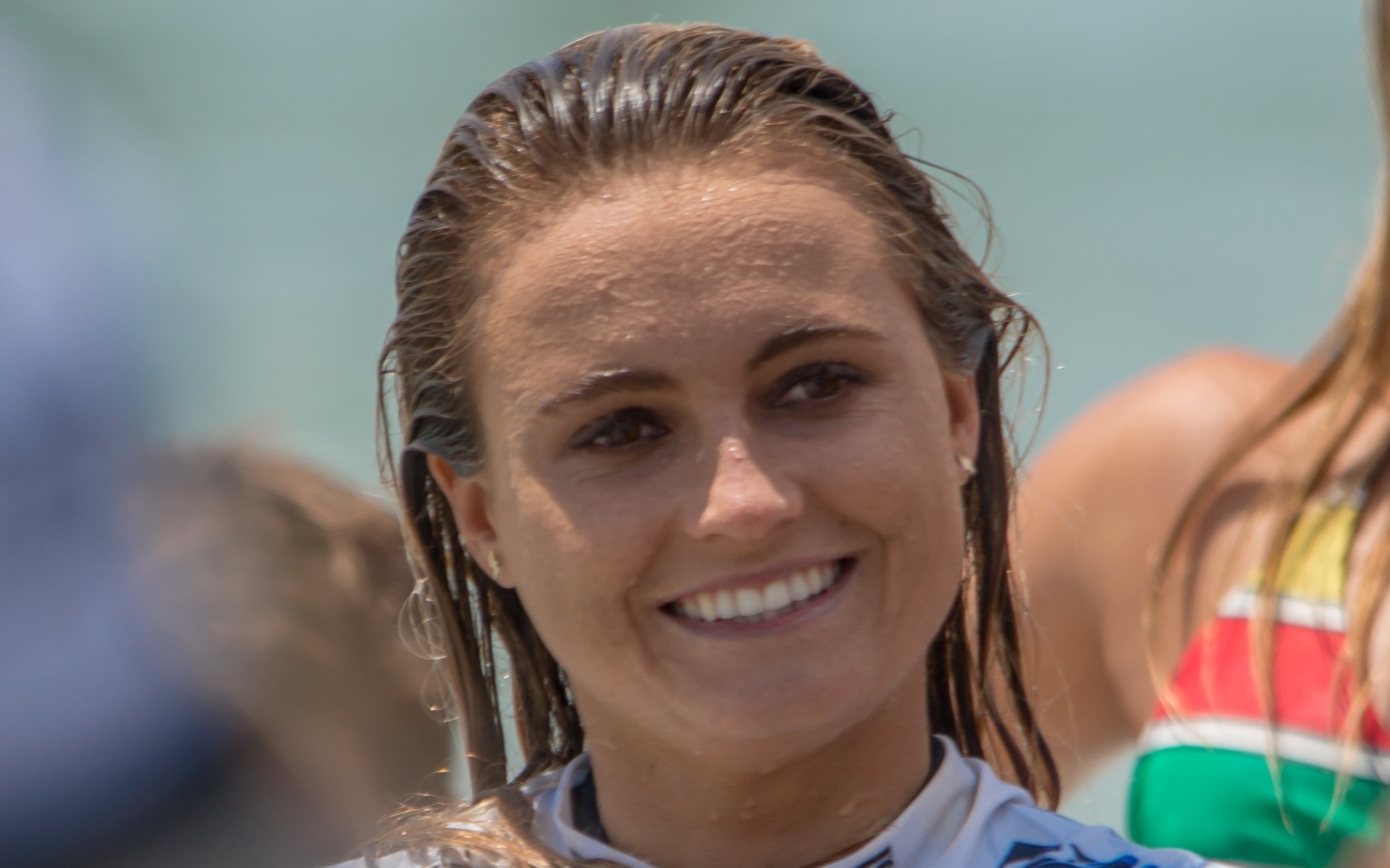 Smiling female surfer in a wetsuit on the beach — sports-themed 2K Quad HD PC desktop wallpaper and background.