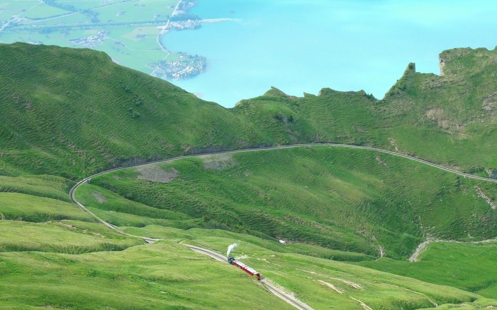 2K Quad HD PC desktop wallpaper: a red steam train vehicle winds along a curving mountain track through lush green hills above a turquoise lake.