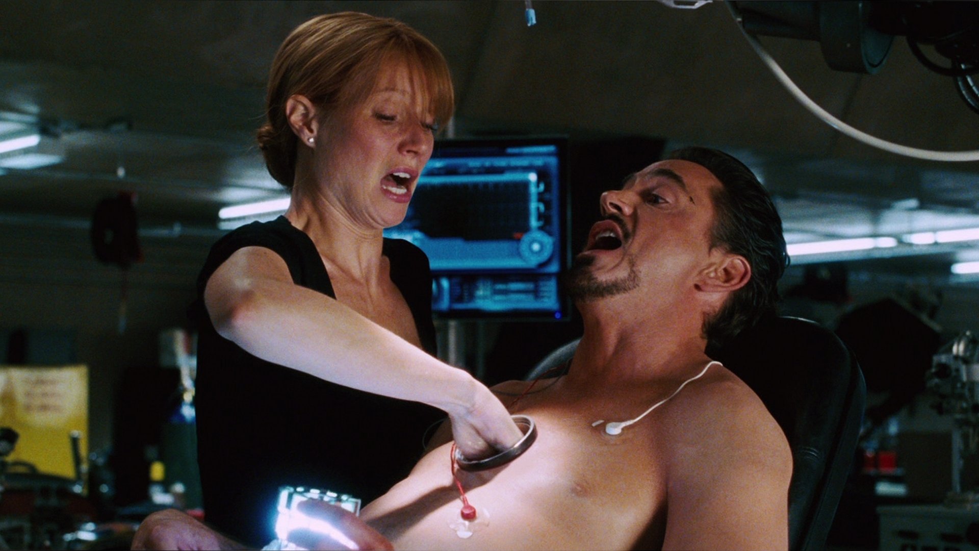 A dramatic scene from Iron Man featuring a woman assisting a man with a device on his chest, set in a high-tech environment. This image serves as an HD desktop wallpaper and background.