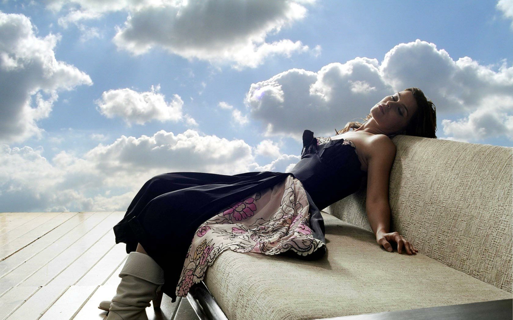 HD PC desktop wallpaper of a female music artist reclining on a modern sofa beneath a bright cloudy sky, wearing a dark dress with a patterned skirt panel.
